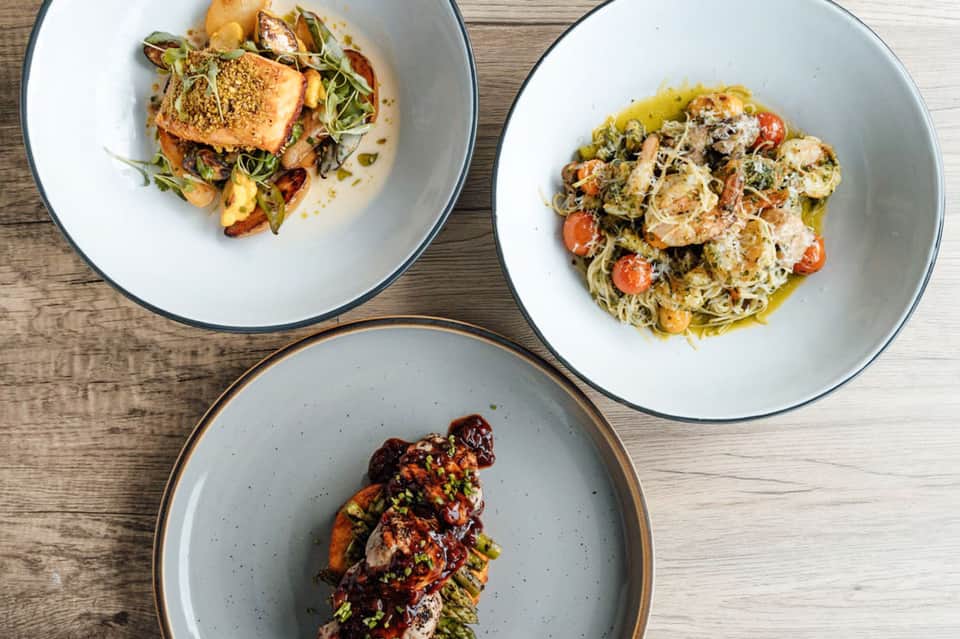 Three plated gourmet seafood dishes on wooden table with fresh fish and vegetable preparations