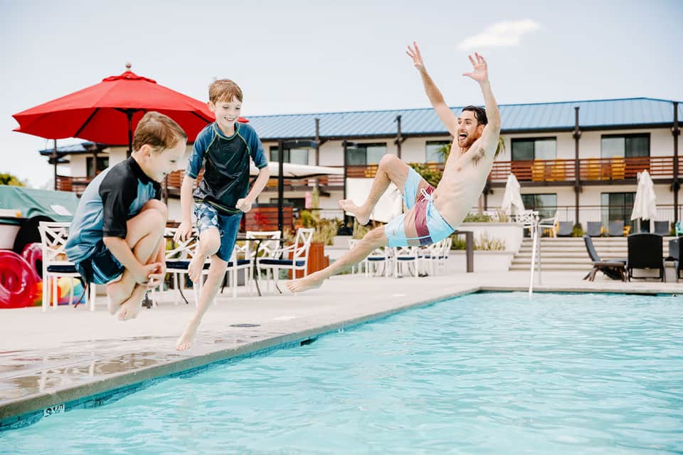Family jumping into resort pool with red umbrella, modern building, and deck seating