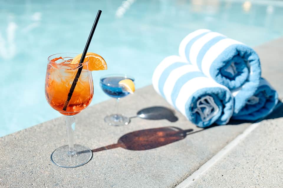 Cocktail with orange slice on pool deck beside rolled blue and white striped towels