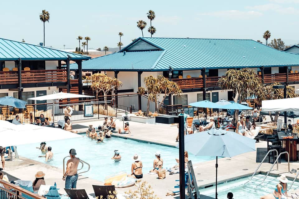 Busy resort pool area with turquoise water, white umbrellas, palm trees, and metal-roofed structures