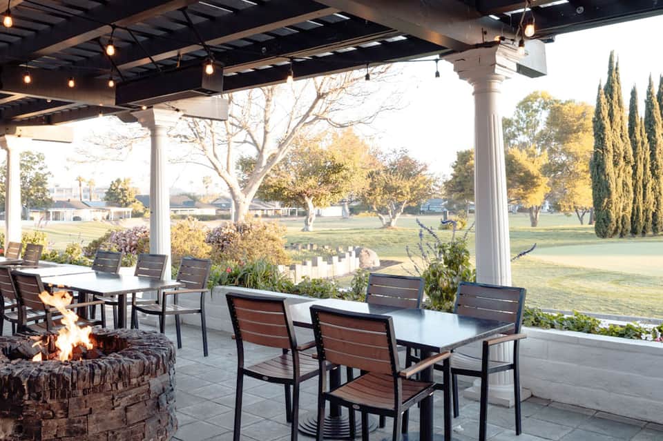 Modern pergola pavilion with dining tables, firepit, and views of a green field and cypress trees