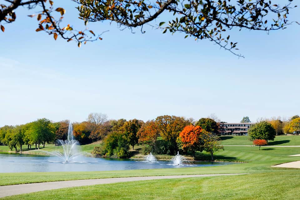 Golf course with manicured lawn, pond with fountains, fall foliage trees, and resort building in distance