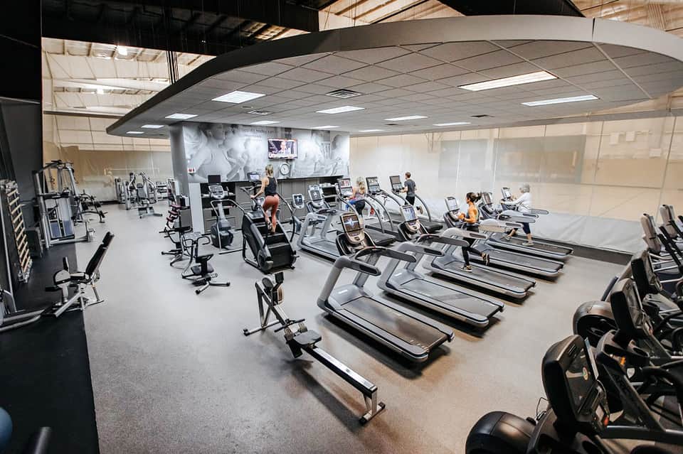 Large fitness studio with rows of treadmills, stationary bikes, and exercise equipment under industrial ceiling