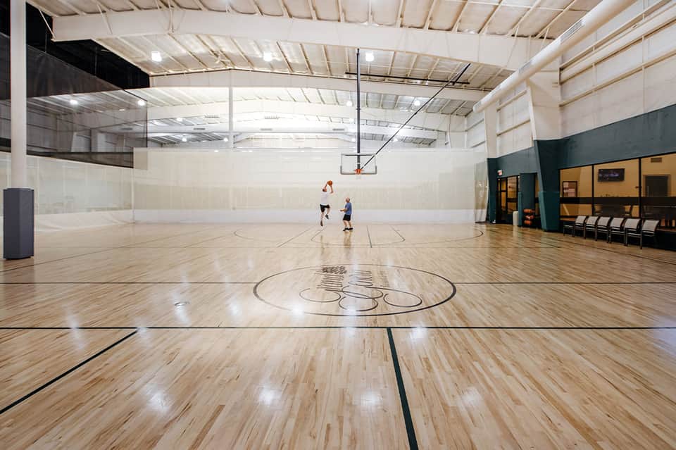 Bright indoor gymnasium with wooden court, basketball hoop, and two players on the floor