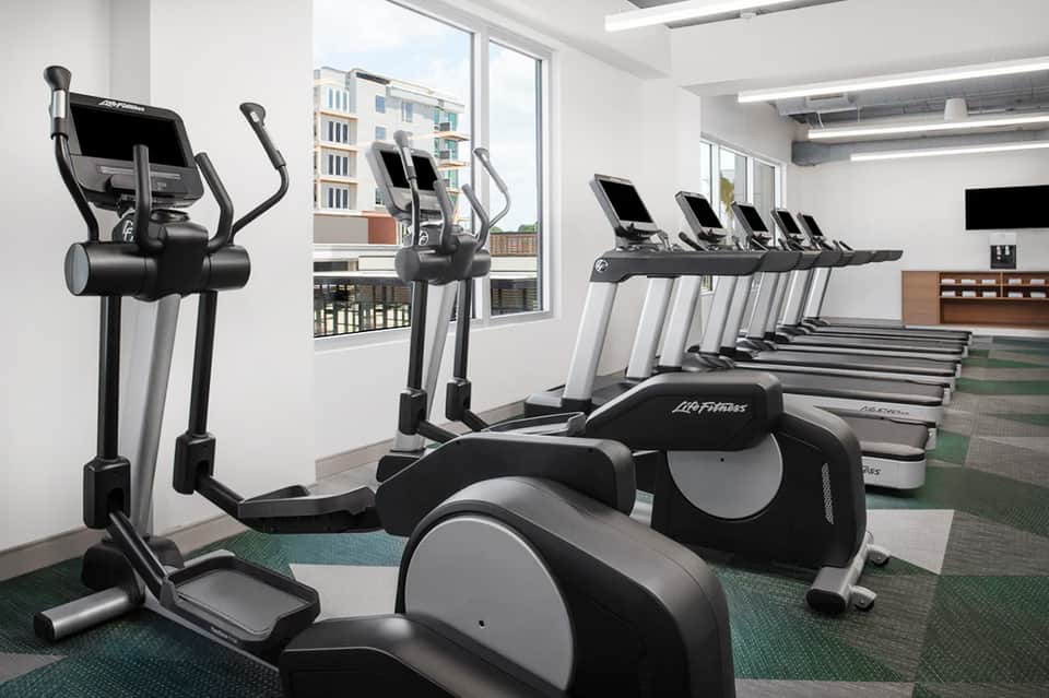 Modern hotel fitness center with elliptical machines, treadmills, large windows overlooking city buildings
