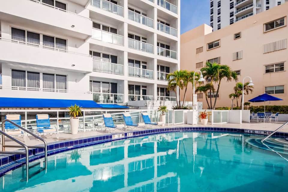 Modern resort pool with blue lounge chairs, white umbrellas, and multi-story buildings
