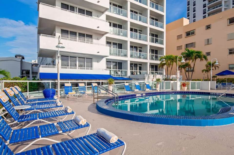 Curved swimming pool with blue lounge chairs beside a high-rise hotel building and palm trees