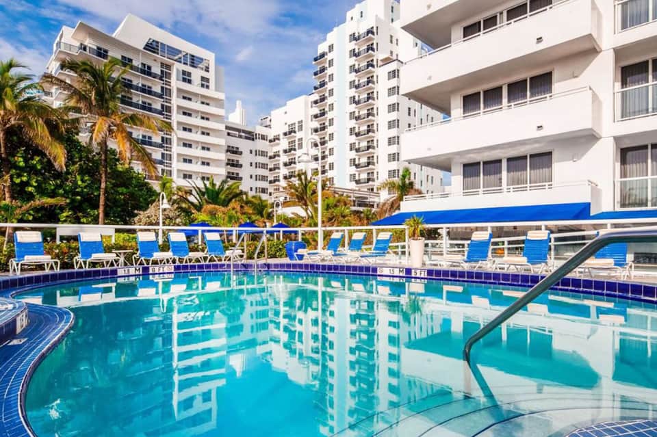 Resort pool with turquoise water, blue lounge chairs, palm trees, and multi-story beachfront hotel buildings