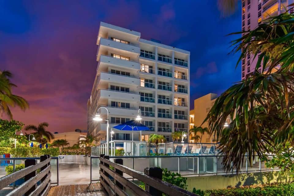Modern waterfront resort building at dusk with lit balconies, palm trees, and blue umbrellas