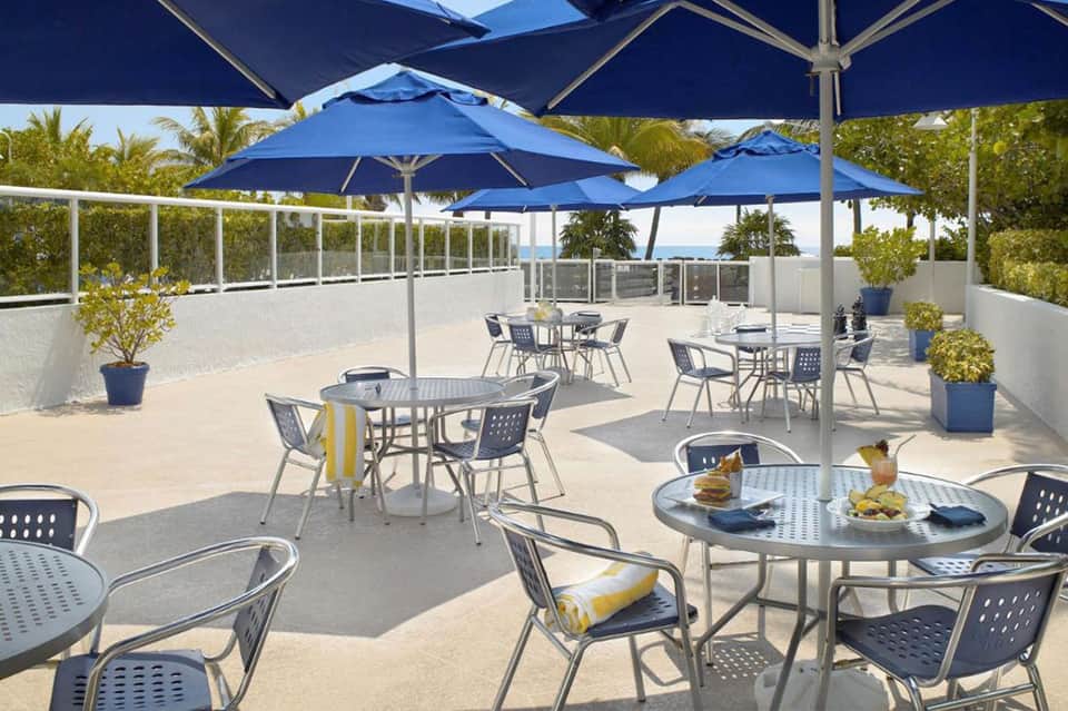 Rooftop terrace with blue umbrellas, metal tables, chairs, white railing, and ocean view