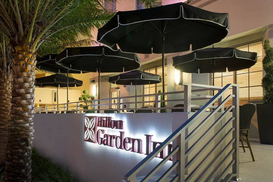 Hilton Garden Inn outdoor terrace with black umbrellas, palm trees, illuminated signage, and evening lighting