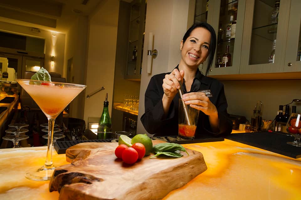 Female bartender preparing cocktails at hotel bar with fresh ingredients and spirits displayed