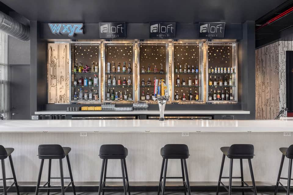 Modern hotel bar with illuminated shelving, black bar stools, and bottle display