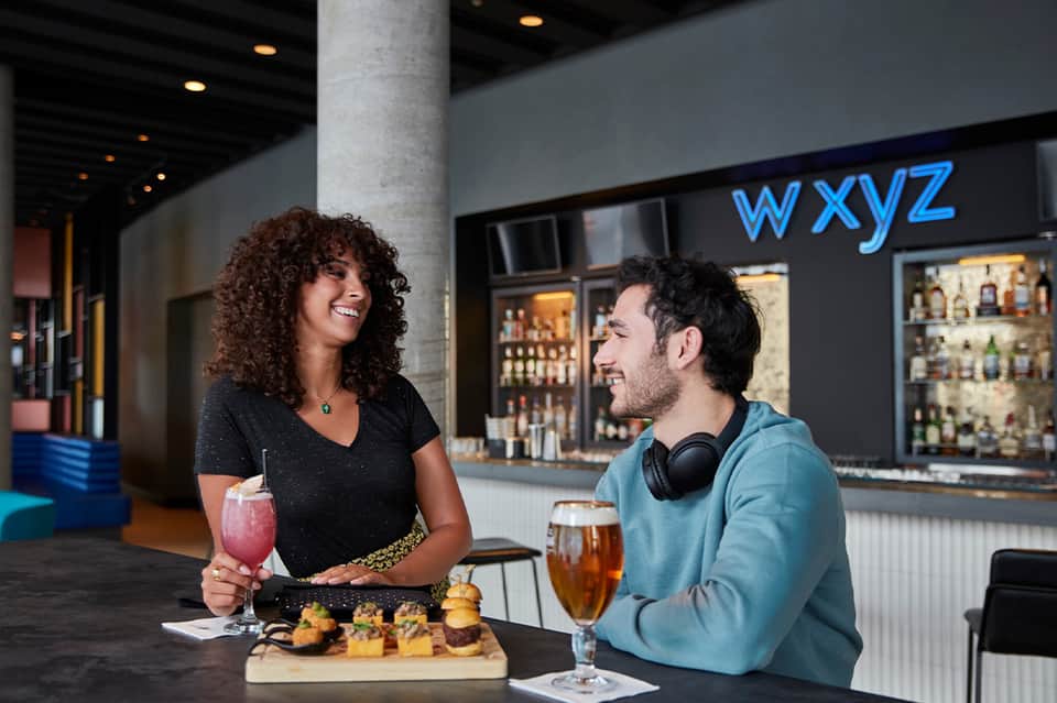 Modern hotel bar interior with neon signage, cocktails, appetizers, and two guests enjoying drinks