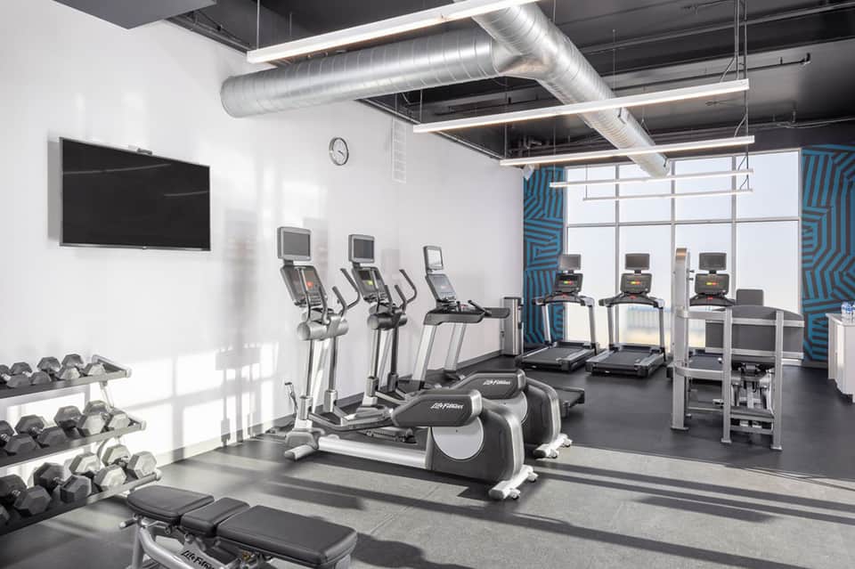 Modern fitness center with cardio equipment, weights, TV screen, and floor-to-ceiling windows