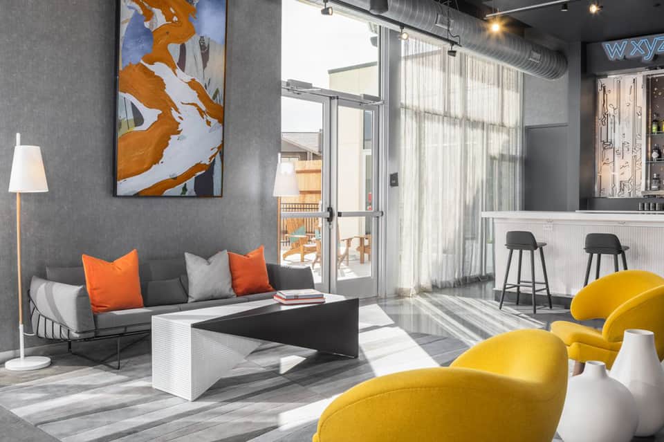 Modern lounge with yellow chairs, gray sofa with orange pillows, abstract art, and glass doors to ocean deck