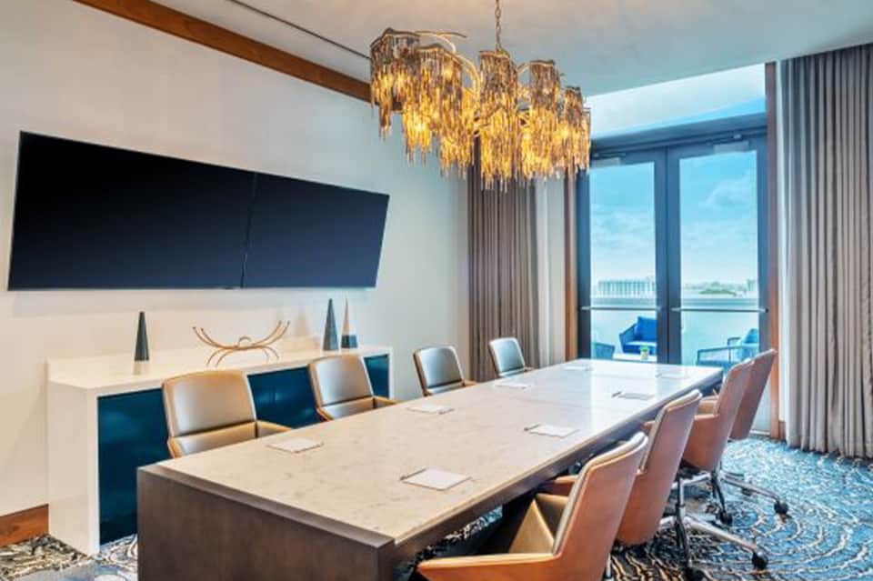 Modern conference room with pale wood table, tan leather chairs, golden chandelier, ocean view terrace