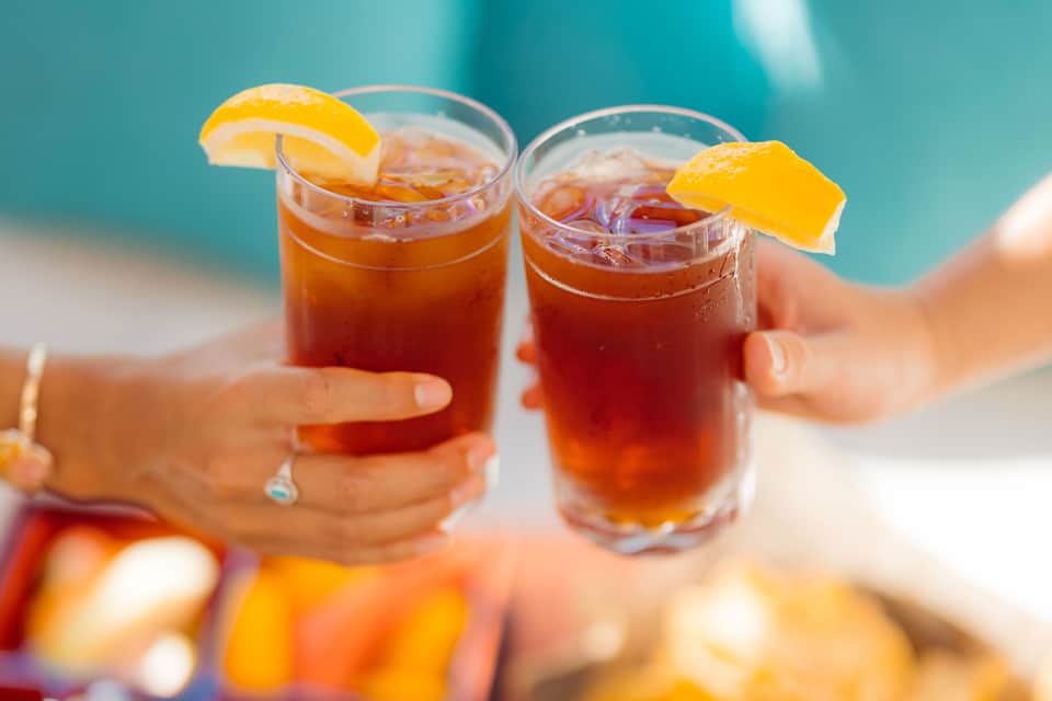 Two hands holding iced beverages with orange slices against beach and ocean background