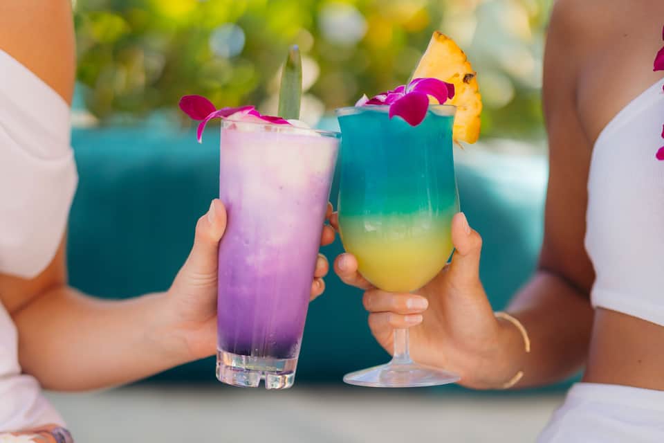 Colorful tropical cocktails with flower garnishes held by people poolside
