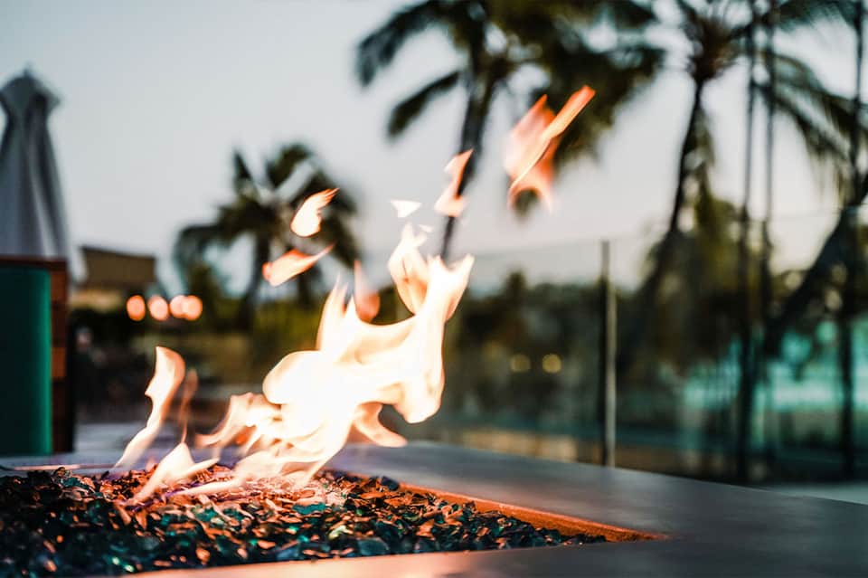 Outdoor fire pit with flames burning, palm trees visible in blurred background at dusk
