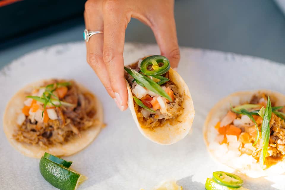 Hand holding gourmet taco with pulled pork, jalapeños, and fresh toppings on white plate