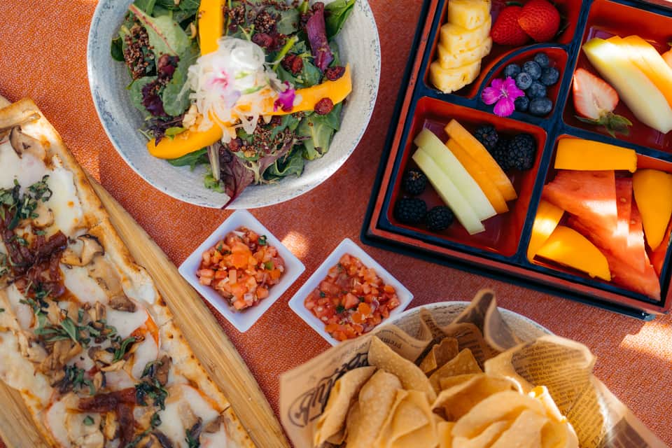 Colorful spread of gourmet food including salad, pizza, sushi, fruit platter, and chips