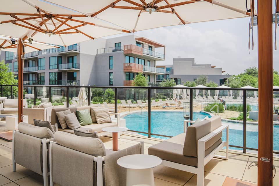 Resort pool terrace with beige lounge seating under umbrellas overlooking modern apartments