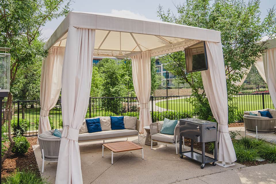 Outdoor lounge pavilion with cream curtains, seating, coffee table, and lush green surroundings