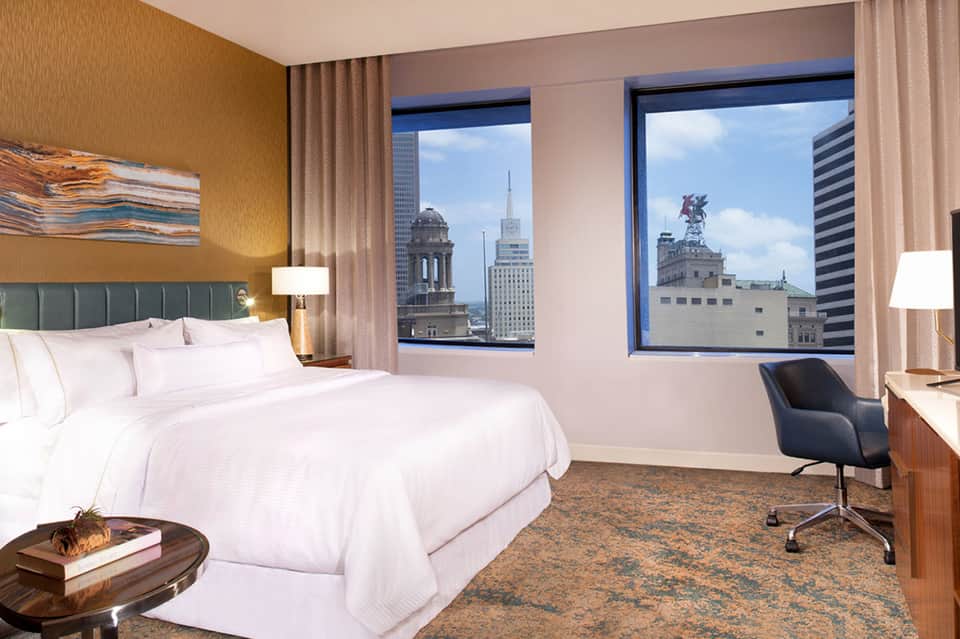 Luxury hotel bedroom with king bed, city skyline views, warm gold walls, and contemporary furnishings