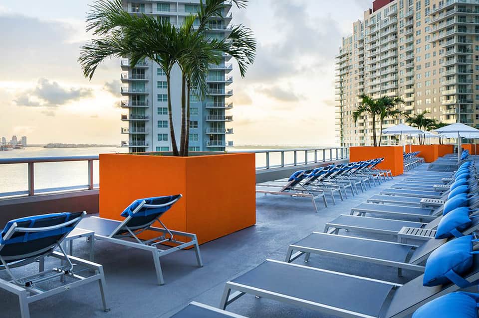 Hotel rooftop pool deck with blue lounge chairs, orange privacy screens, and waterfront skyline