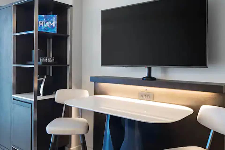 Modern hotel room workspace with large TV, white desk, and black shelving unit