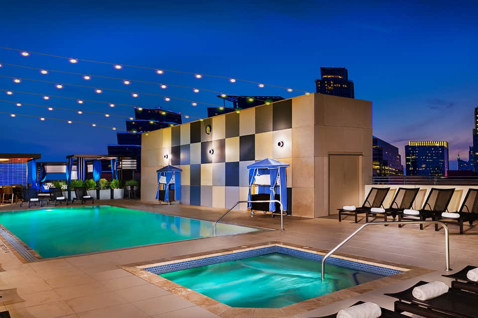 Rooftop pool and hot tub with city skyline at dusk, string lights overhead, and modern architecture