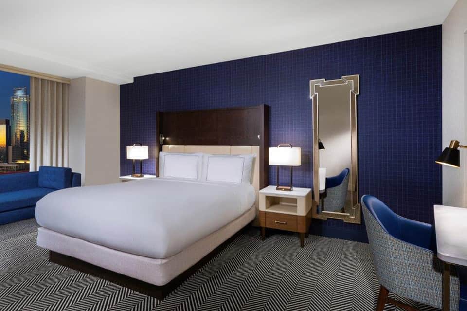Hotel bedroom with navy accent wall, king bed, blue seating, and city views through windows