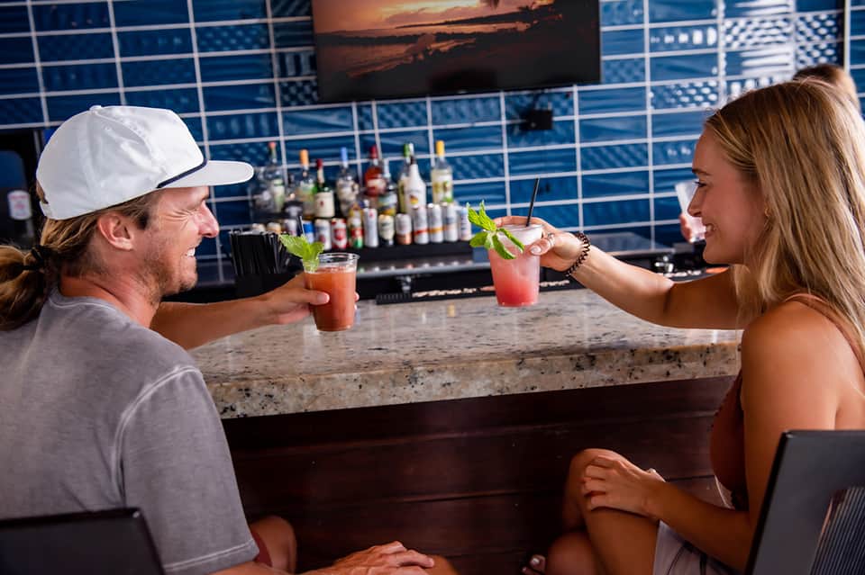 Two guests enjoying cocktails at a rooftop bar with ocean backdrop and well-stocked liquor shelves