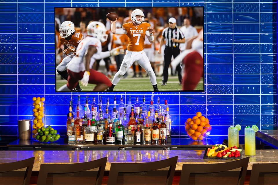 Sports bar with large screen showing Texas football game, blue tile wall, and liquor bottles display
