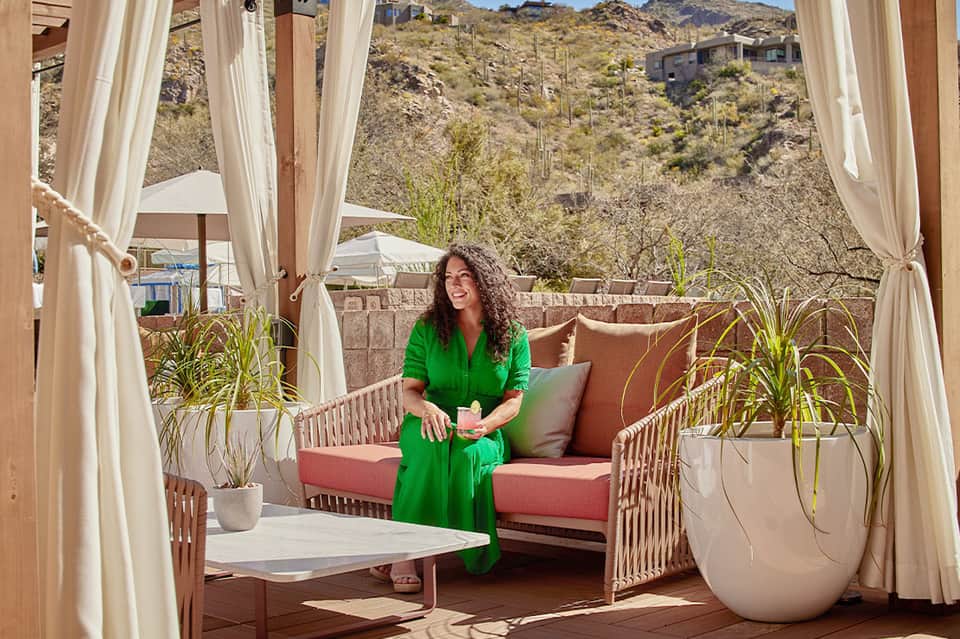 Woman in green dress sitting on coral cushioned seating with desert mountain views and curtains
