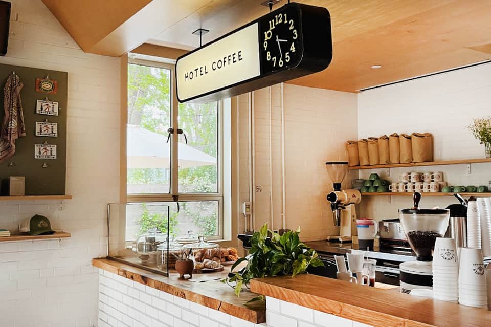 Hotel coffee station with vintage clock sign, wooden counter, shelving with supplies, and green plants