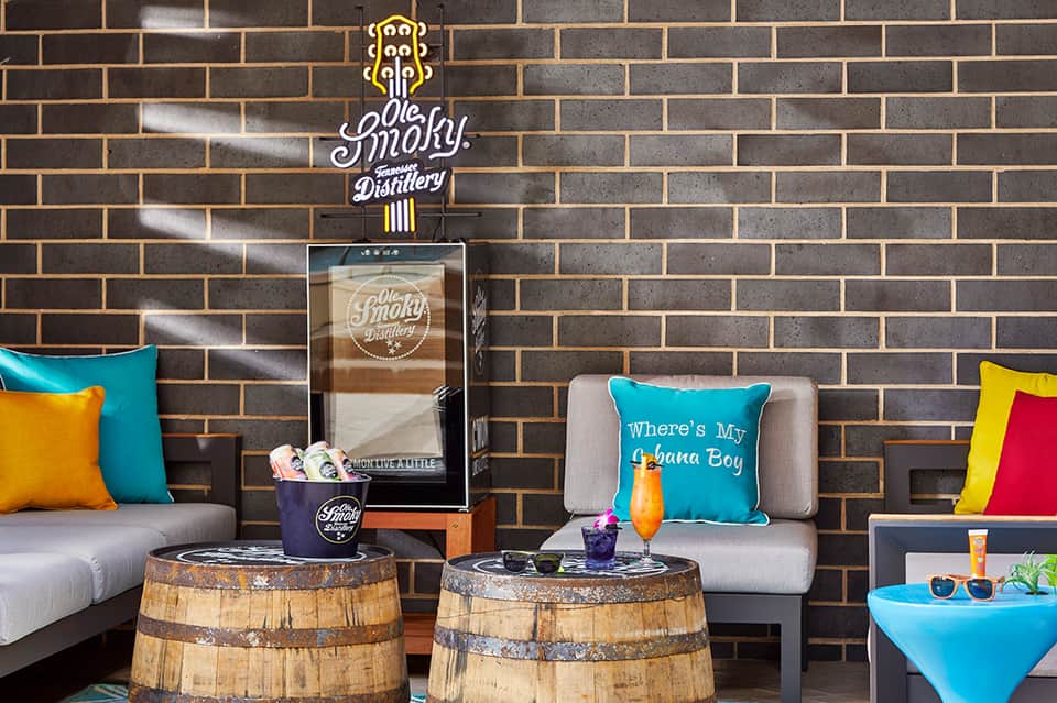 Modern lounge area with colorful pillows, brick wall, barrel tables, and distillery sign