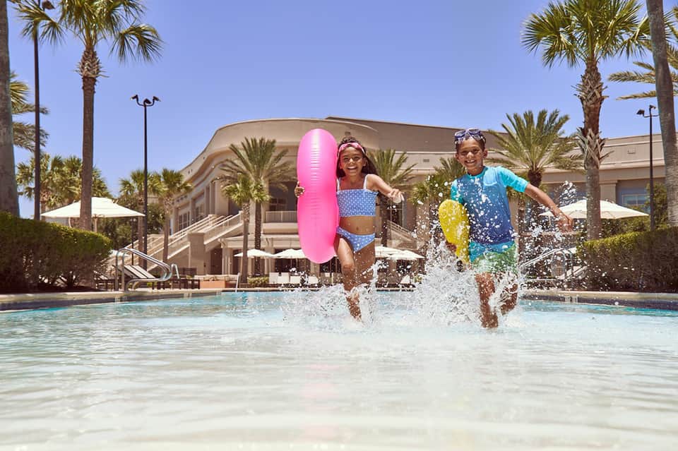 Children running with inflatable floats into shallow resort pool with palm trees
