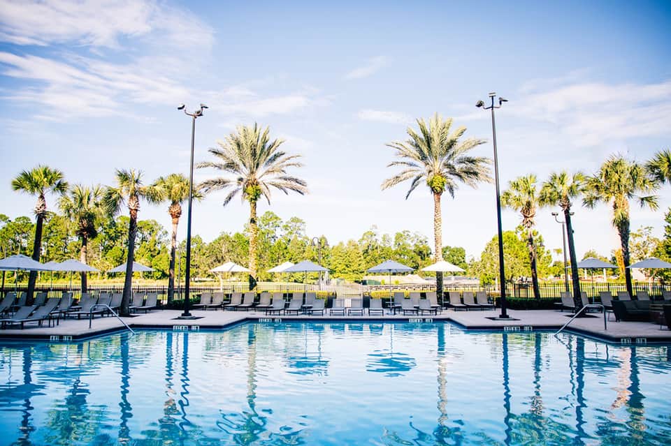 Resort swimming pool with palm trees, lounge chairs, and umbrellas under blue sky