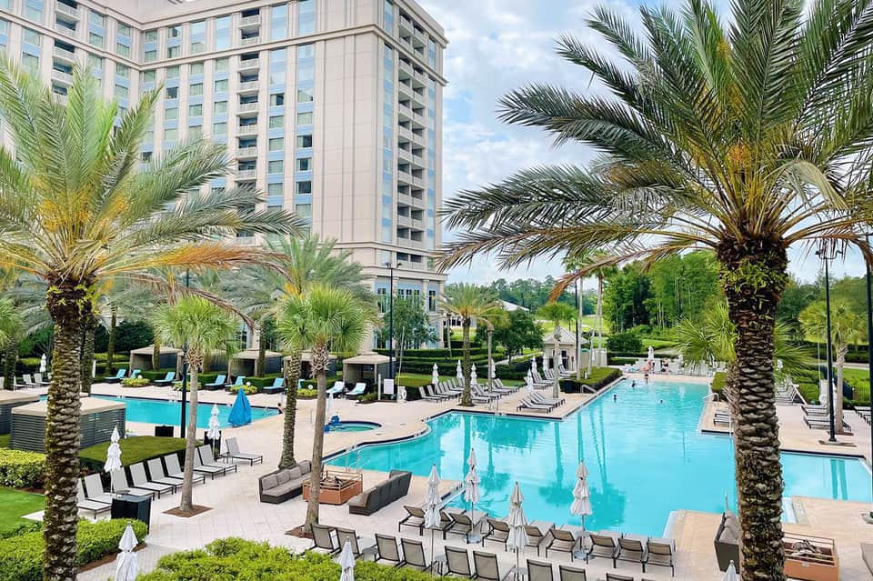 Large resort pool with palm trees, lounge chairs, hotel tower, and manicured landscaping