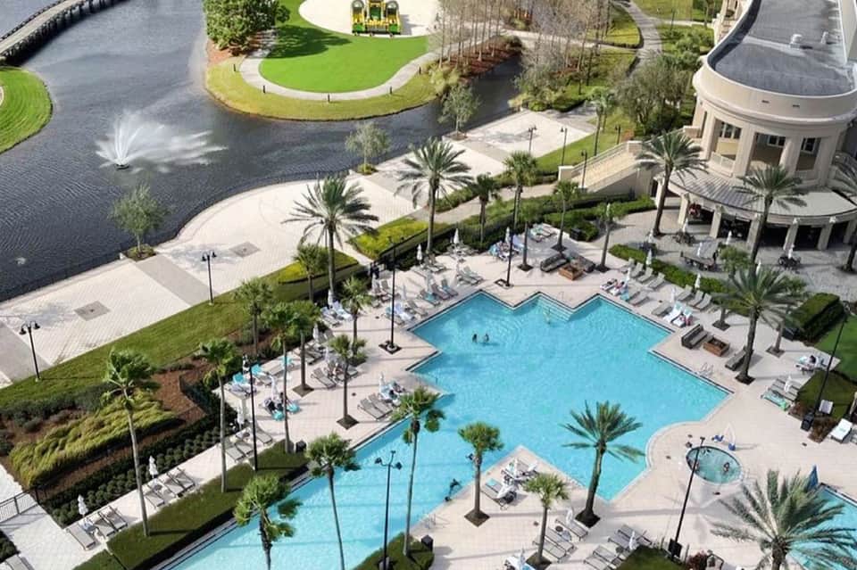 Aerial view of resort pool complex with palm trees, buildings, and manicured grounds near waterway