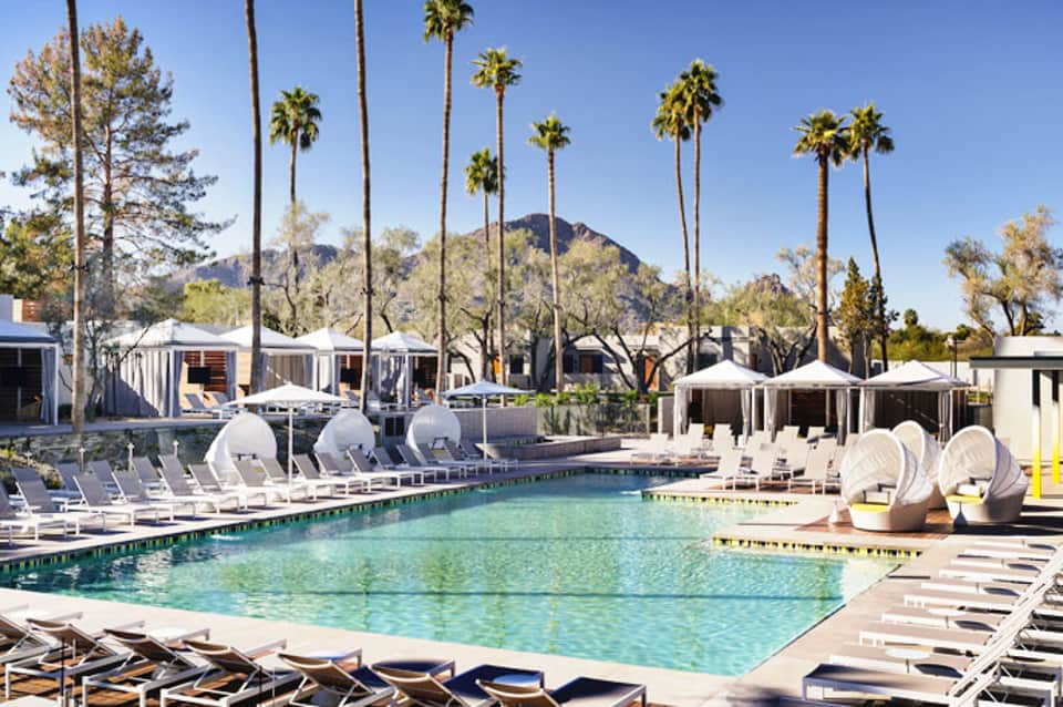 Resort pool with palm trees, lounge chairs, cabanas, and mountain backdrop under clear blue sky