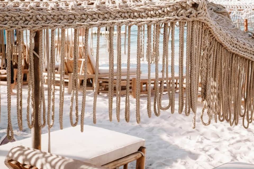 Beachside cabana with macramé fringe canopy, white cushioned seating, and ocean view through ropes