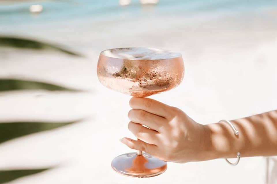 Hand holding rosé cocktail with frosted glass against blurred beach background