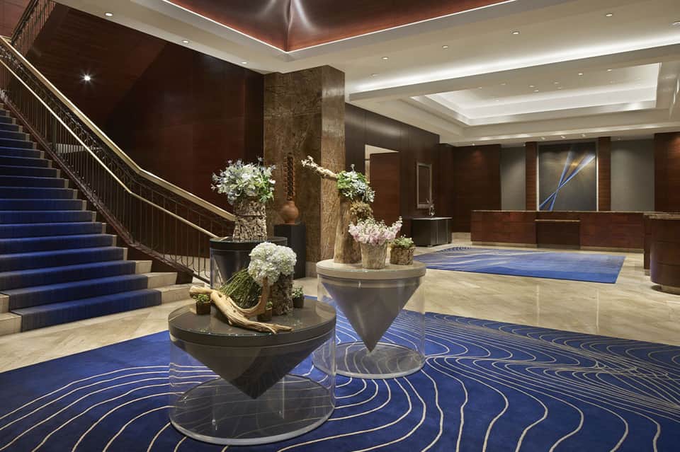 Luxury hotel lobby with blue-patterned carpet, curved staircase, and sculptural floral arrangements