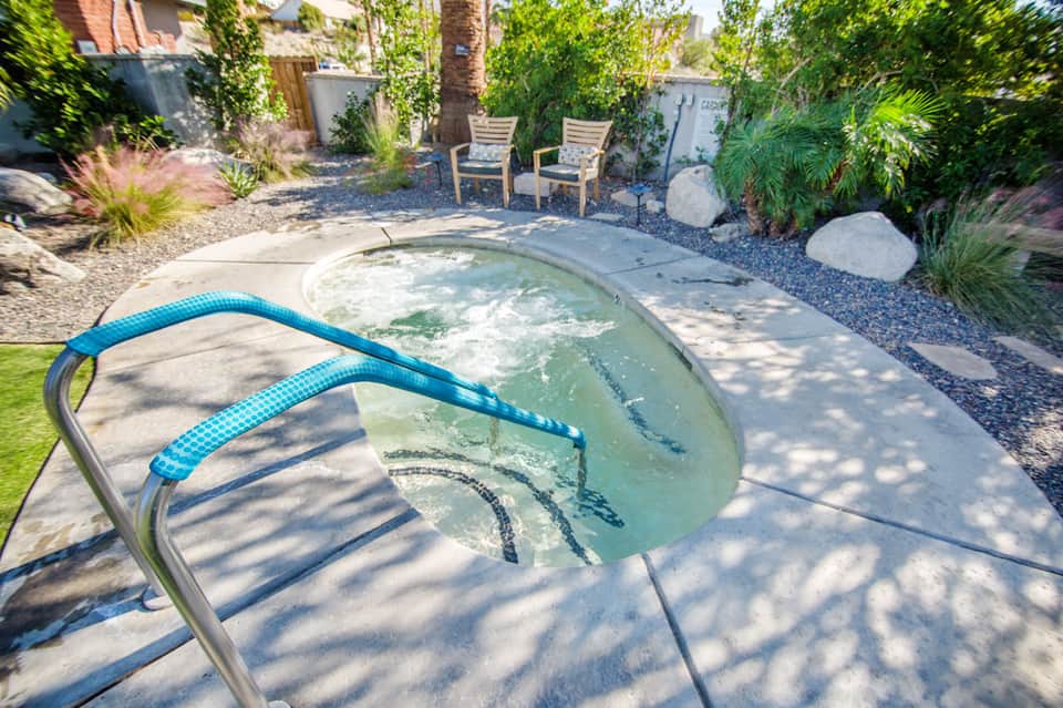 Neglected hot tub with algae and blue cleaning hose in landscaped backyard with palm trees