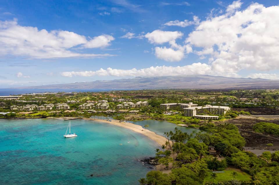 Aerial resort view of turquoise bay with sailboat, sandy beach, and distant volcanic mountain