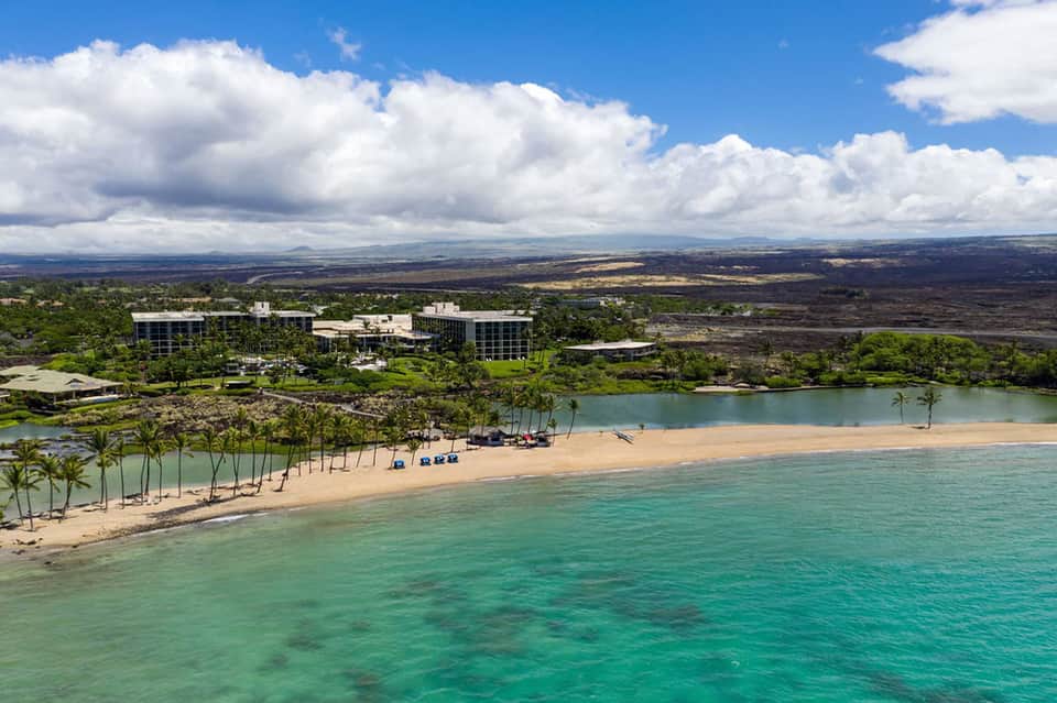 Aerial beachfront resort with sandy shore, lagoon, multi-story buildings, and surrounding lush landscape