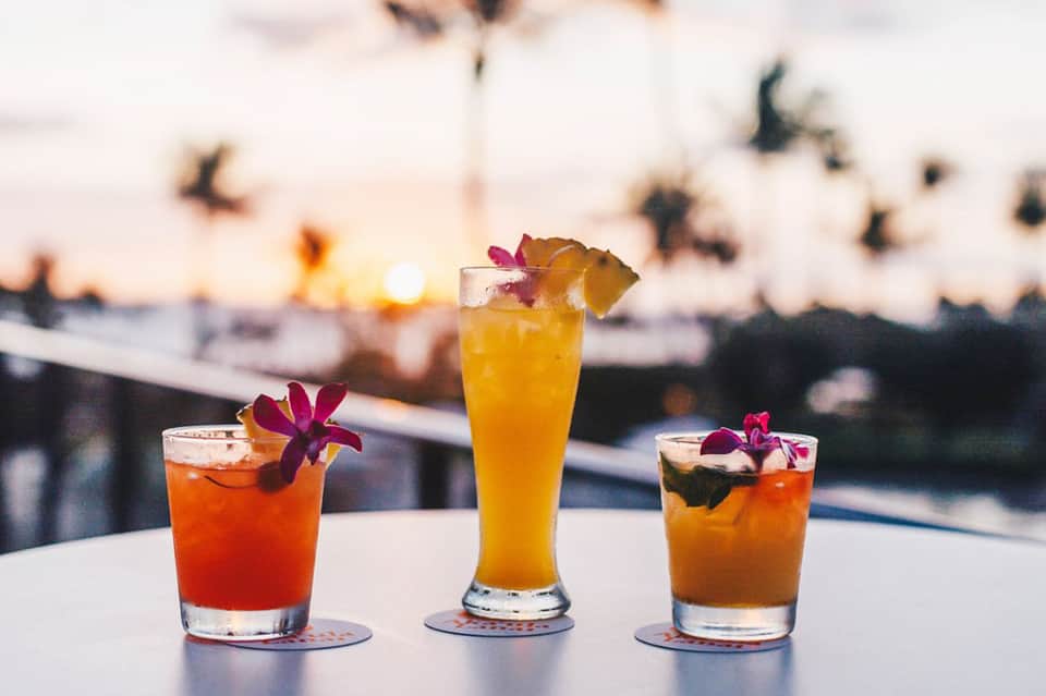Three tropical cocktails with colorful flowers at oceanfront bar during golden hour